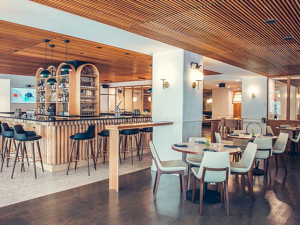 The image shows a modern, stylish bar area with high stools around the bar and several dining tables with chairs in an elegantly designed interior.