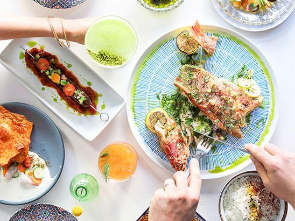 A table with various dishes: grilled fish, appetizers, rice, and colorful drinks. Two people are seen reaching for the food and drinks.