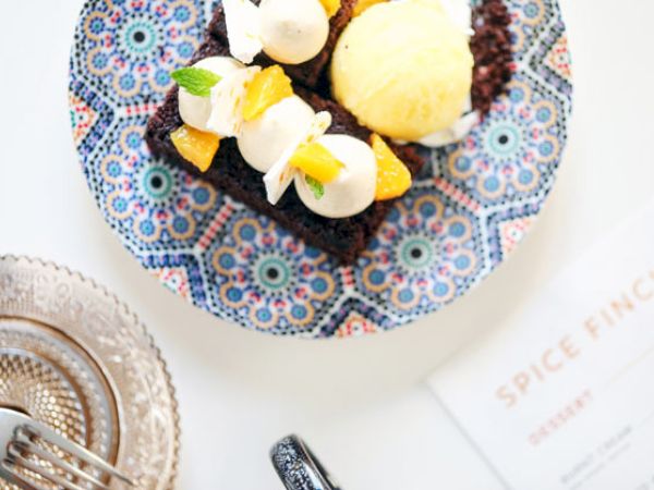 A dessert plate with cake topped with cream, fruit, and ice cream, next to cutlery and a glass plate. A "Spice Finch" menu is partially visible.