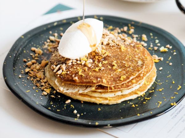The image shows a plate of pancakes topped with whipped cream, syrup being poured, and sprinkled nuts and zest on a dark plate.