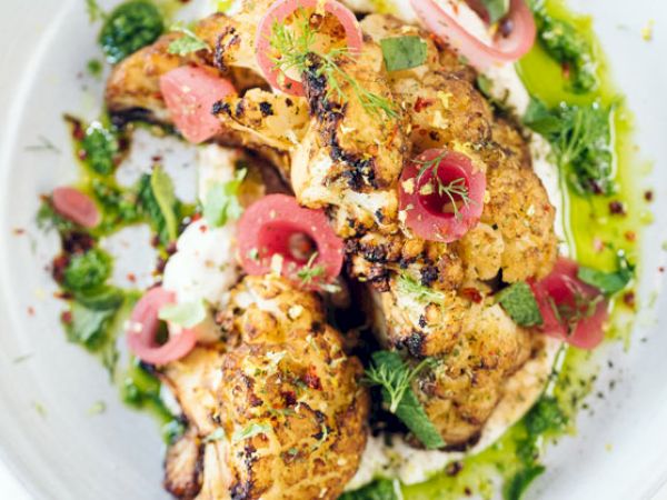 The image shows a beautifully plated dish featuring roasted cauliflower topped with pickled red onions and garnished with fresh herbs and a green sauce.