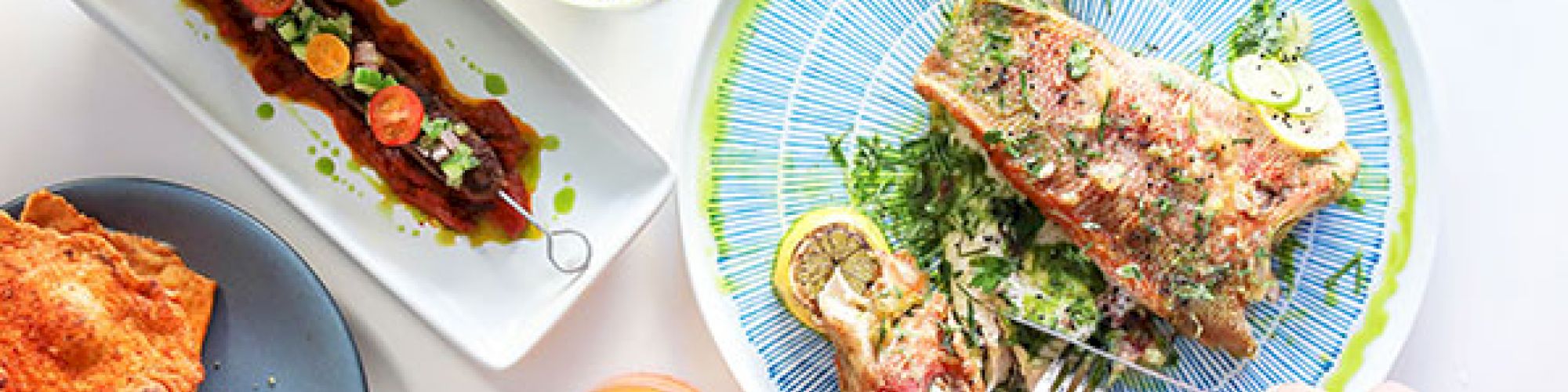 A table set with various dishes, including fish, appetizers, salads, rice, and drinks, with people reaching for food.