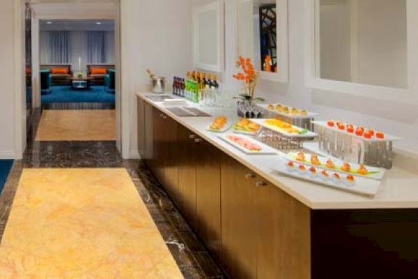 The image shows a reception area with a table, chairs, a food and beverage counter, and a coffee machine. There is a hallway in the background.