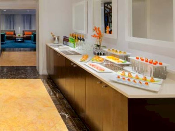 The image shows a reception area with a table, chairs, a food and beverage counter, and a coffee machine. There is a hallway in the background.