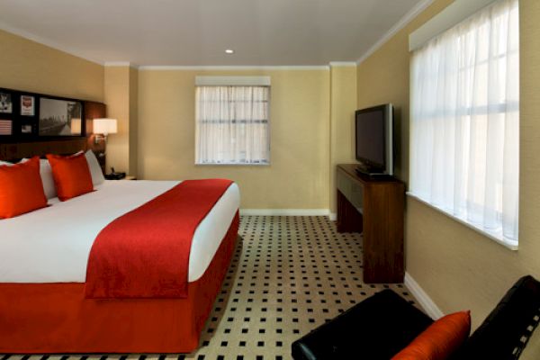 A cozy hotel room featuring a large bed with red accents, a flat-screen TV, and two large windows covered with sheer white curtains.