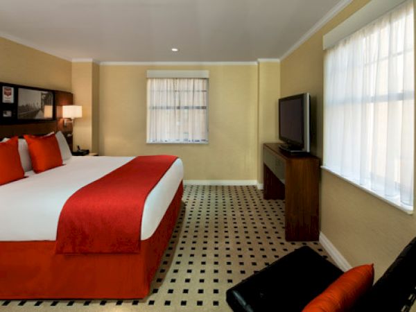 A cozy hotel room featuring a large bed with red accents, a flat-screen TV, and two large windows covered with sheer white curtains.