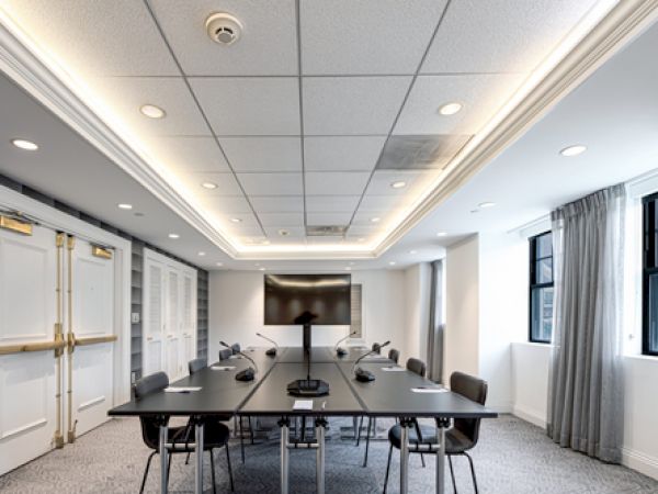 A modern conference room with a long black table, chairs, a large screen monitor, and microphones. The room has recessed lighting and windows.