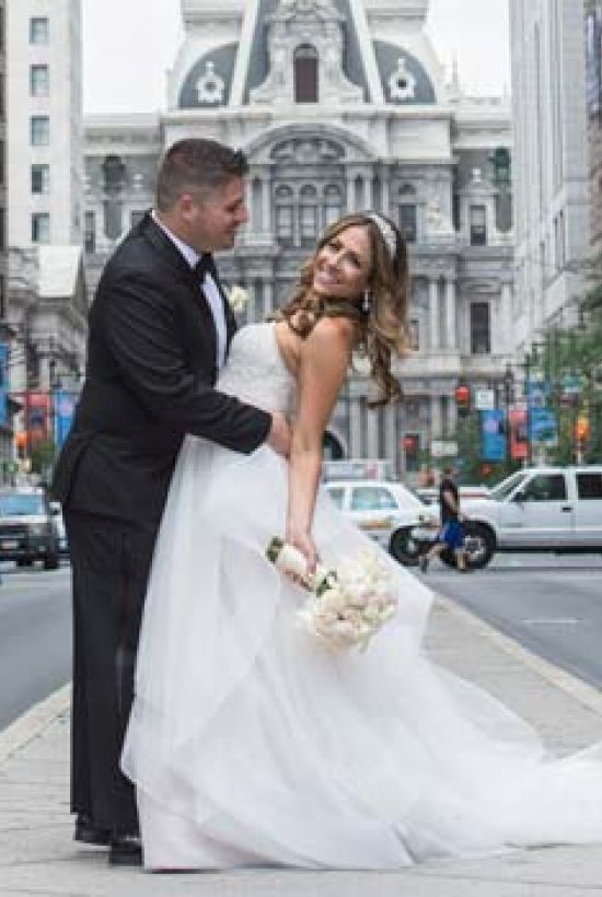 A couple dressed in wedding attire poses in the middle of a city street, with the city hall in the background and traffic around them.