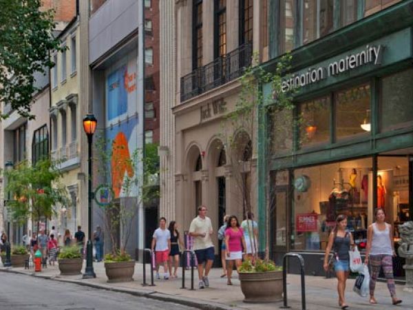 People are walking along a city sidewalk past various shops, including "destination maternity." Streetlamps and planters line the street.