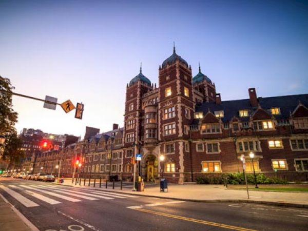 A large, historic building, likely part of a university or institution, is seen at dusk with street lights and a crosswalk in the foreground.
