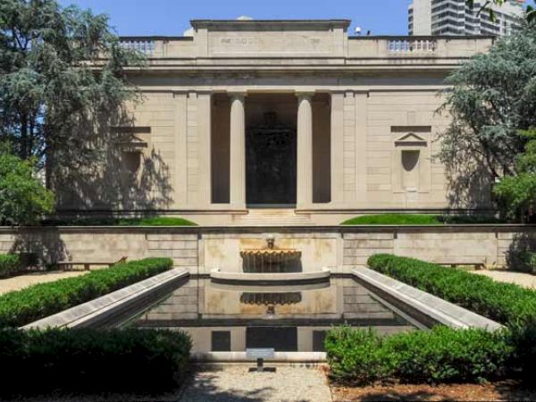 The image shows a neoclassical building with pillars at the entrance, surrounded by greenery, and a reflecting pool in front of it.