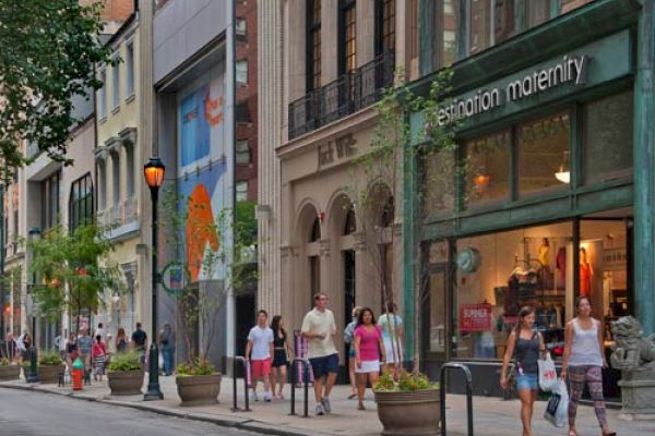 Pedestrians walk along a city sidewalk, passing by store fronts including a maternity shop, with trees and plants lining the street.