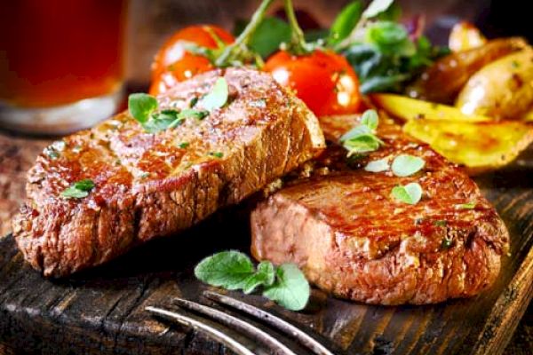 The image shows two grilled steaks topped with herbs, accompanied by cherry tomatoes and roasted potatoes on a wooden cutting board.