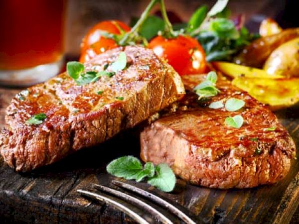 The image shows two grilled steaks topped with herbs, accompanied by cherry tomatoes and roasted potatoes on a wooden cutting board.