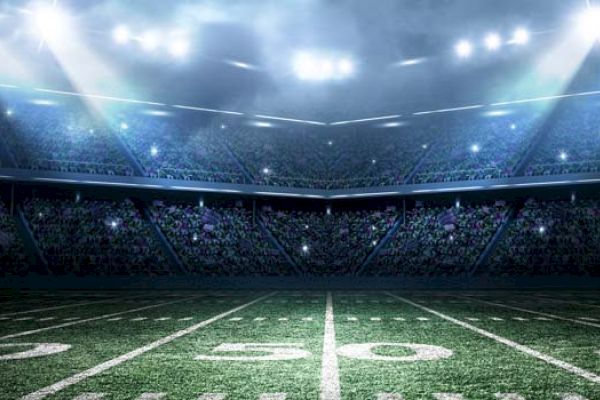 A well-lit football stadium at night, showing the field's 50-yard line and filled stands under bright stadium lights.