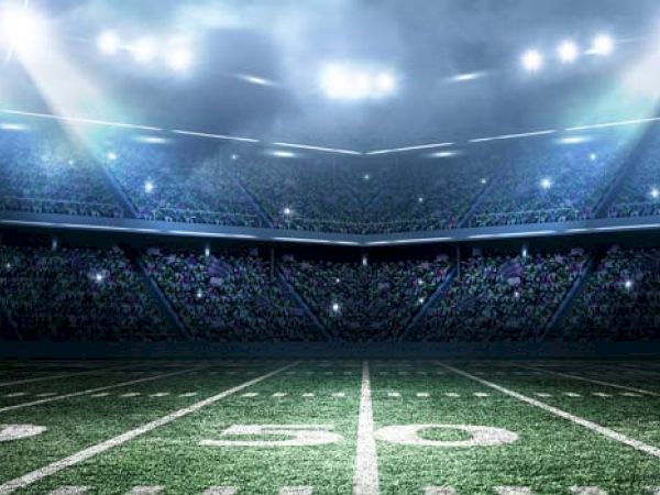 A well-lit football stadium at night, showing the field's 50-yard line and filled stands under bright stadium lights.