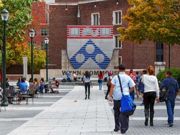 People are walking and sitting near a building with a large University of Pennsylvania logo and a sign reading "Wynn Commons" in the background.