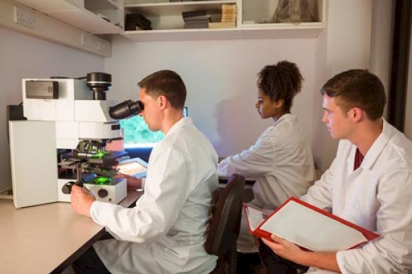 Three scientists in lab coats are conducting research, with one looking into a microscope and the other two taking notes in a laboratory setting.