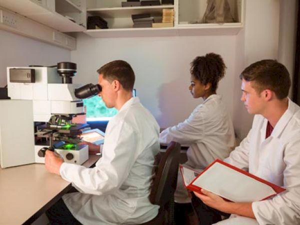 Three scientists in lab coats are conducting research, with one looking into a microscope and the other two taking notes in a laboratory setting.