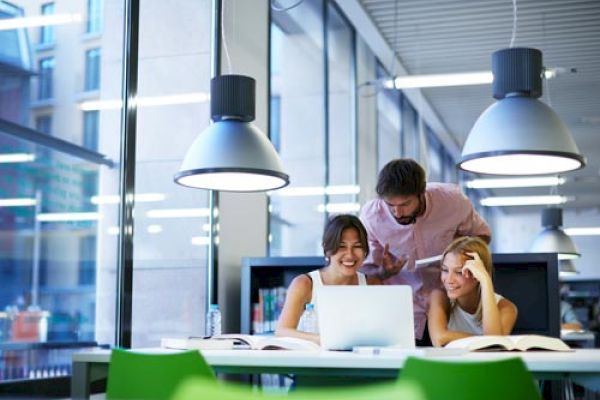 Three people are gathered around a laptop in a bright, modern setting. They appear to be engaged in a discussion or study session.