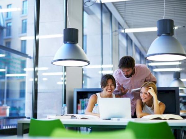 Three people are gathered around a laptop in a bright, modern setting. They appear to be engaged in a discussion or study session.