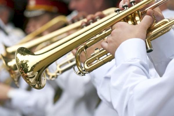 The image shows a close-up of brass trumpet players wearing white uniforms, possibly part of a marching band or musical group.