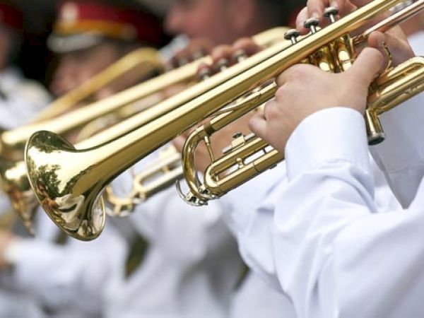 The image shows a close-up of brass trumpet players wearing white uniforms, possibly part of a marching band or musical group.