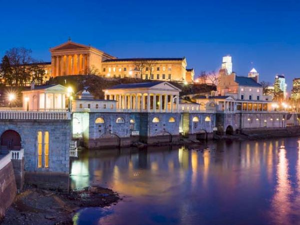 The image shows the Philadelphia Museum of Art and the Fairmount Water Works in Philadelphia, beautifully lit up at dusk with city skyscrapers in the background.