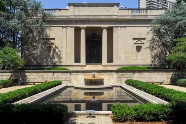 This image shows a classical-style building with columns, manicured gardens, and a reflective pool in front. The scene is tranquil and symmetrical.