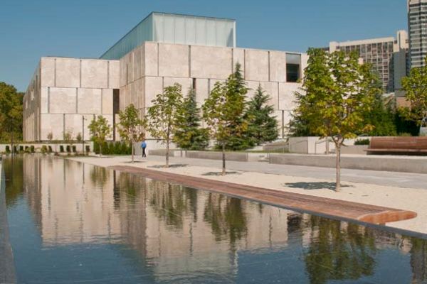 This image shows a modern building with a reflective pond in front, surrounded by trees and a paved walkway under a clear blue sky.