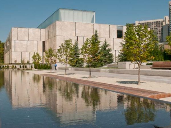 This image shows a modern building with a reflective pond in front, surrounded by trees and a paved walkway under a clear blue sky.