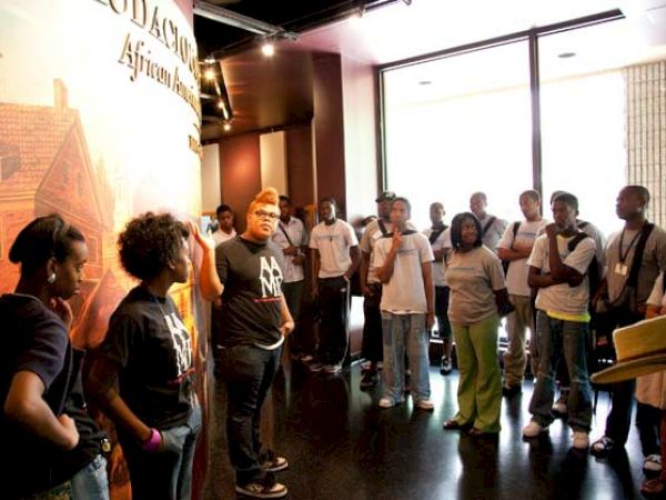 A group of people is gathered indoors, listening to a person speaking near a display. They appear to be on a tour or in an educational setting.