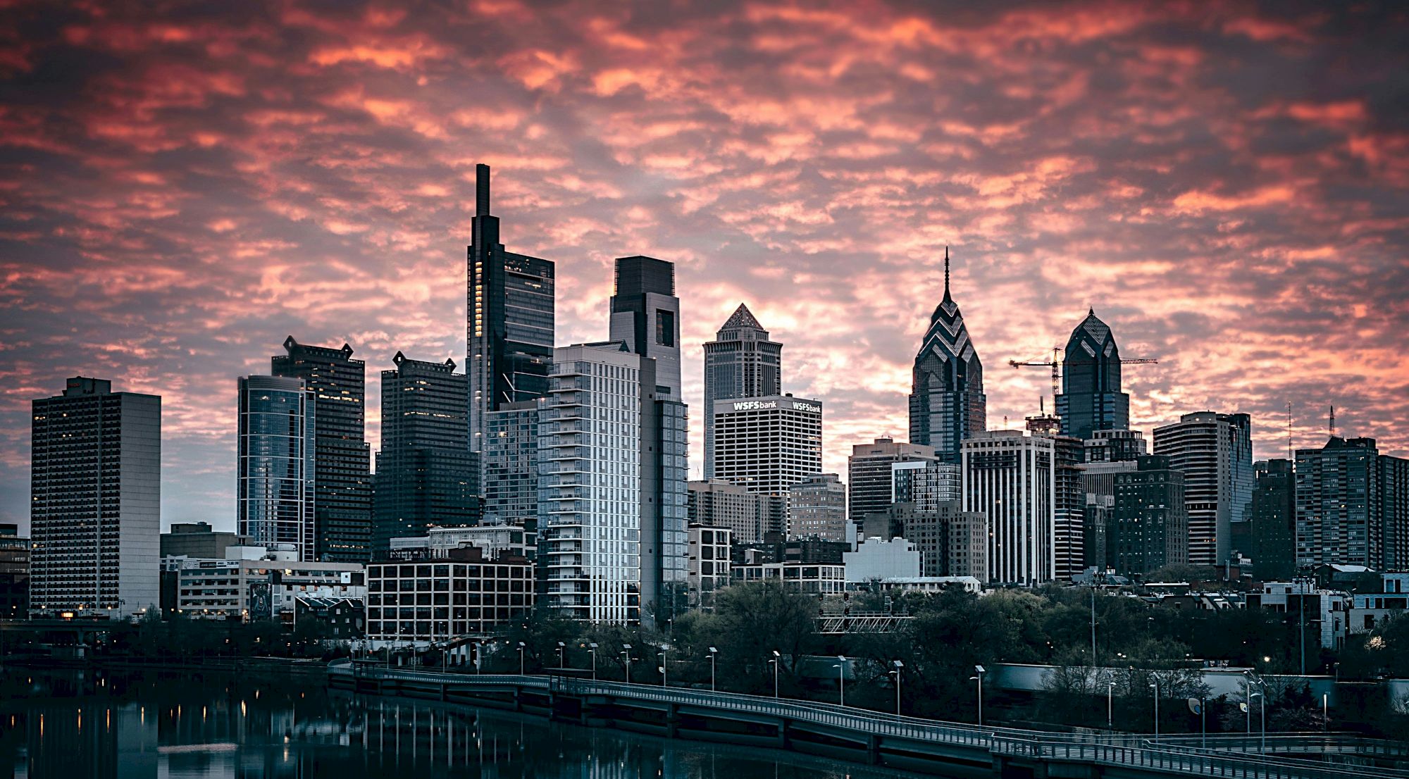 The image shows a city skyline at sunset with a mixture of modern and classic skyscrapers, under a dramatic, cloud-filled sky reflected in a body of water.