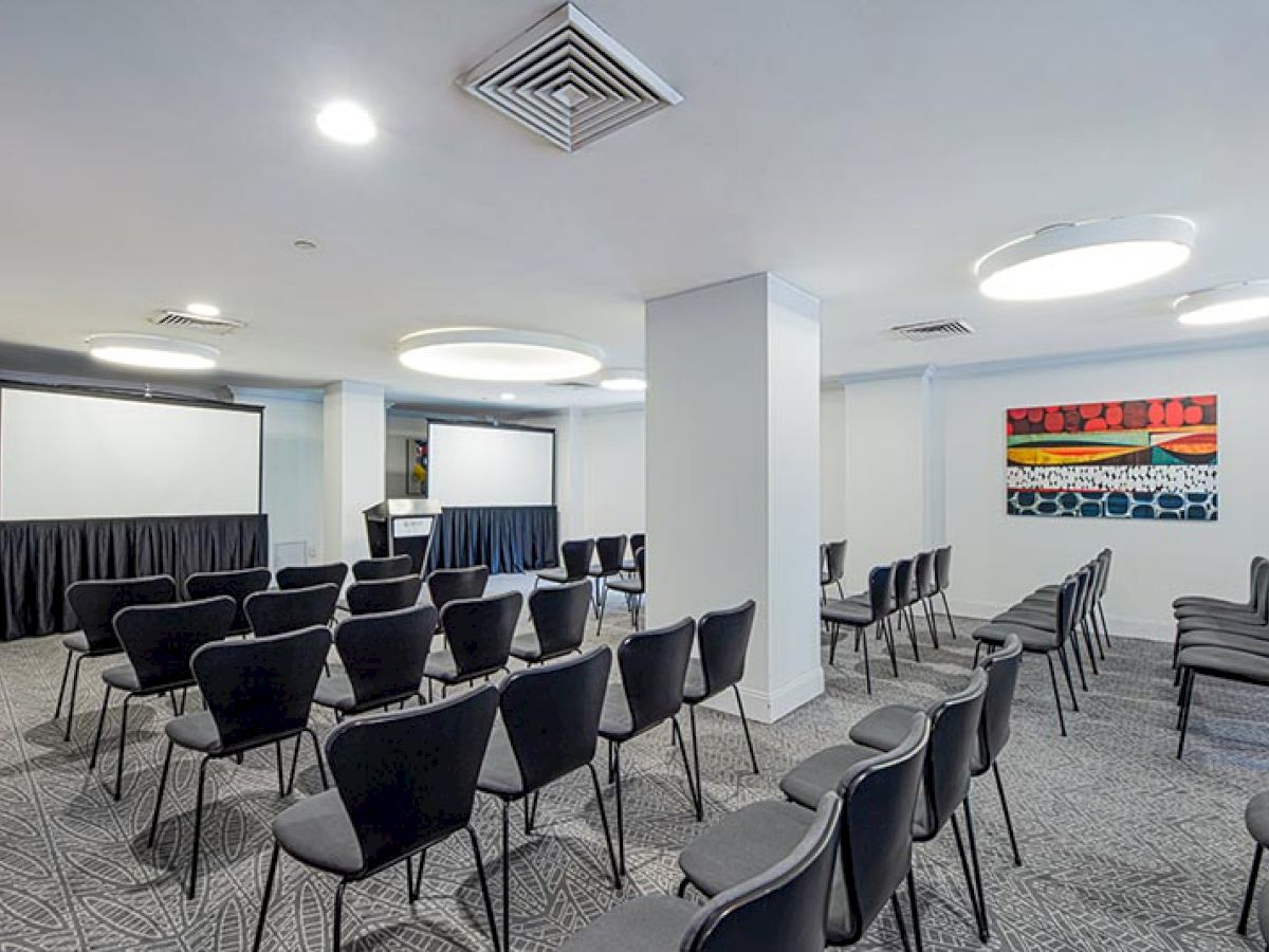 The image shows a modern, well-lit conference or presentation room set up with rows of chairs facing two projection screens.