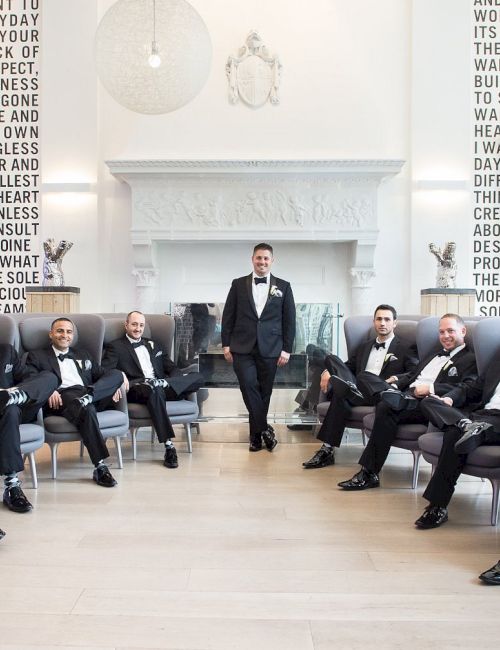 A group of men in tuxedos sit and stand in a well-lit room, with large text covering the walls behind them.