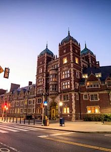 A large, historic building with illuminated windows is seen at twilight, flanked by a street with a crosswalk and traffic lights.