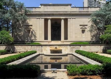 A classical building fronted by a symmetrical garden with a rectangular reflective pool, trimmed hedges, and a decorative central fountain.