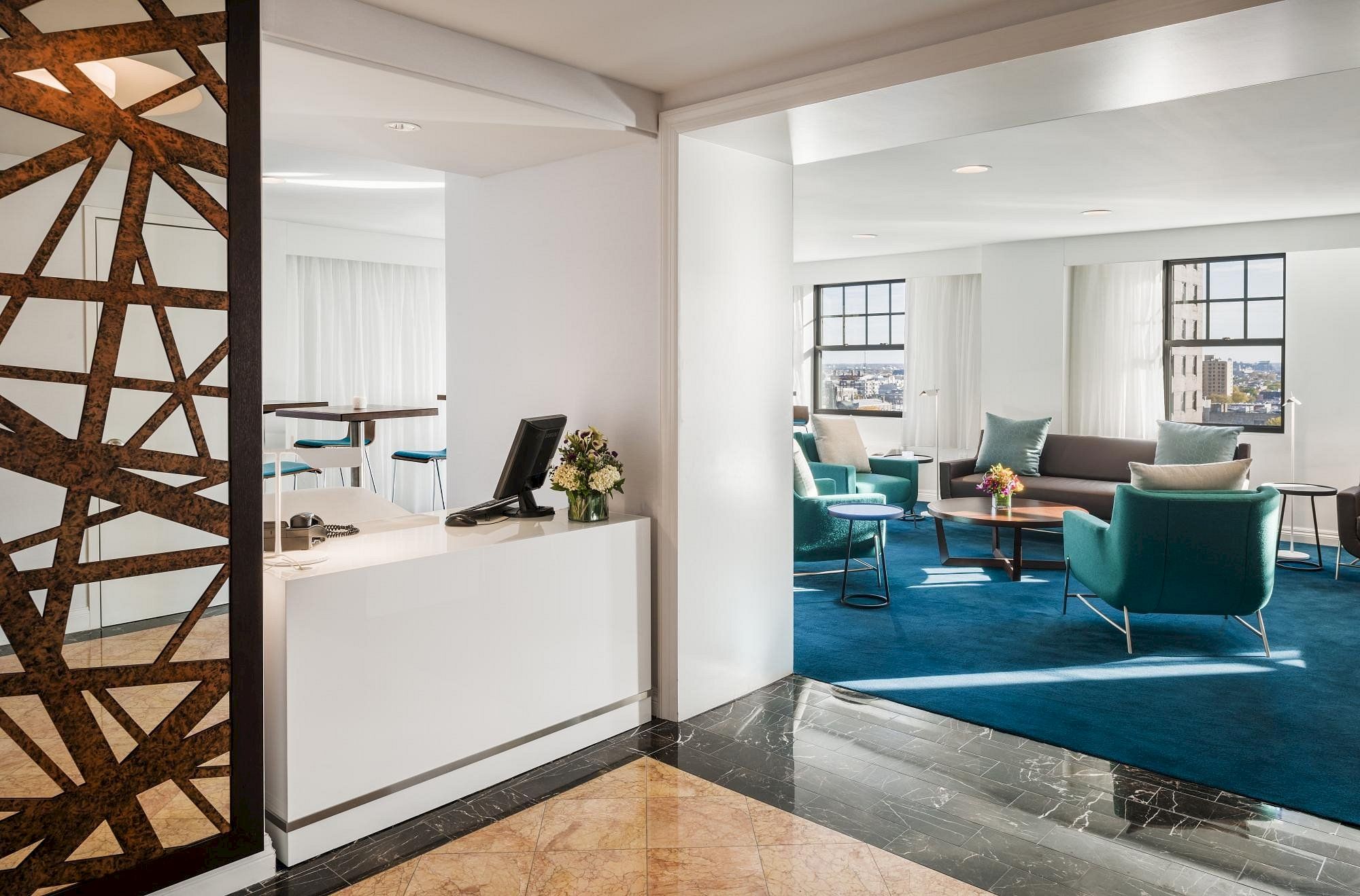 A modern living area with blue carpet, teal chairs, sofas, and windows. The entrance has a small desk, wooden decor, and flower centerpiece.