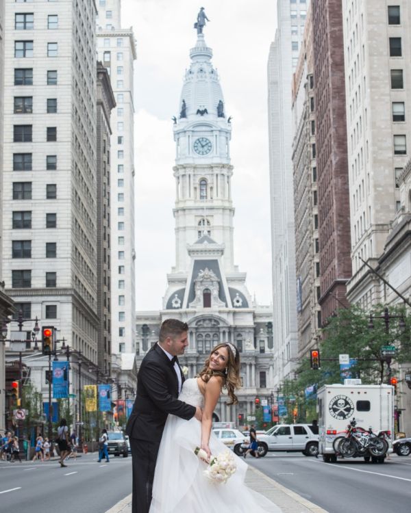 A couple in wedding attire poses in the middle of a city street with a tall, historic building in the background amidst skyscrapers and vehicles passing by.