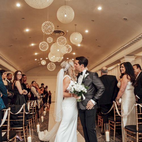 A bride and groom kiss at the end of the wedding aisle, surrounded by guests in a beautifully decorated indoor venue.