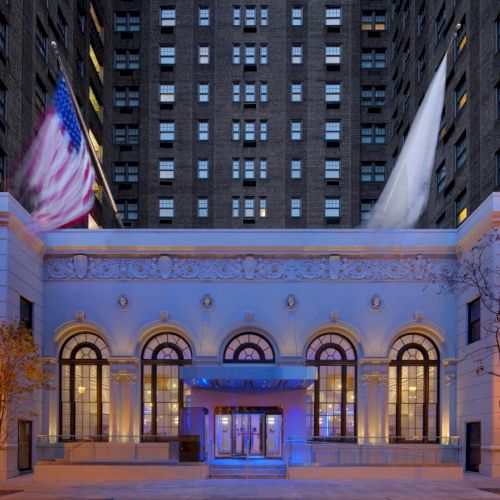 An elegant building entrance with large windows, decorative details, and flags, flanked by tall buildings, illuminated in soft light.