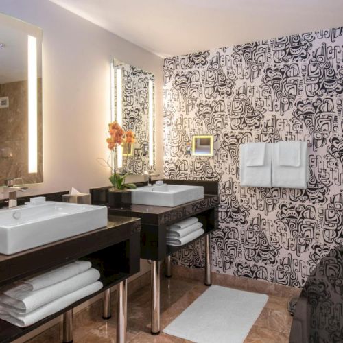 A modern bathroom with two sinks, a bathtub, a patterned accent wall, and towels. It includes mirrors with lighting and some decorative items.