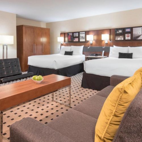 A hotel room with two beds, a couch, yellow pillows, modern chairs, a coffee table with green apples, lamps, and a wardrobe.