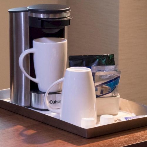 A coffee maker on a tray with two white mugs, coffee packets, creamer packs, and a few other small items is placed on a wooden surface.