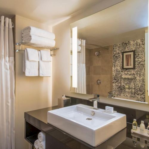 A modern bathroom with a large illuminated mirror, a square sink, towel rack with folded towels, shower with curtain, and various toiletries.