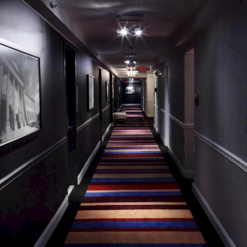 A dimly lit, long corridor with a colorful striped carpet and framed pictures on the walls under ceiling lights, creating a cozy ambiance.