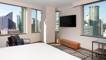 The image depicts a modern hotel room with large windows offering a city skyline view, a wall-mounted TV, and minimalistic furniture.