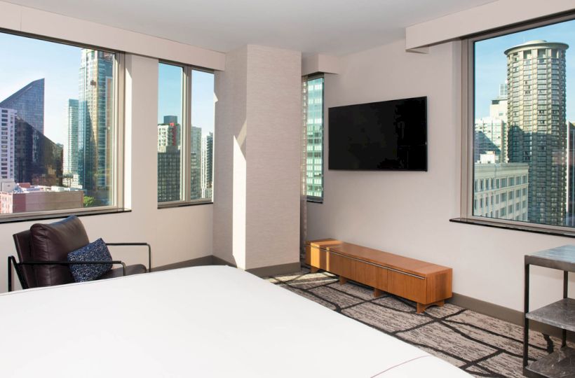 The image depicts a modern hotel room with large windows offering a city skyline view, a wall-mounted TV, and minimalistic furniture.