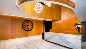 A modern reception area with a marble desk, a large wall clock, wooden paneling, a spiral staircase, and a flower arrangement.