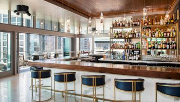 The image shows a modern, well-lit bar interior with a sleek design. There are chairs with blue cushions, a fully stocked bar, and large windows.